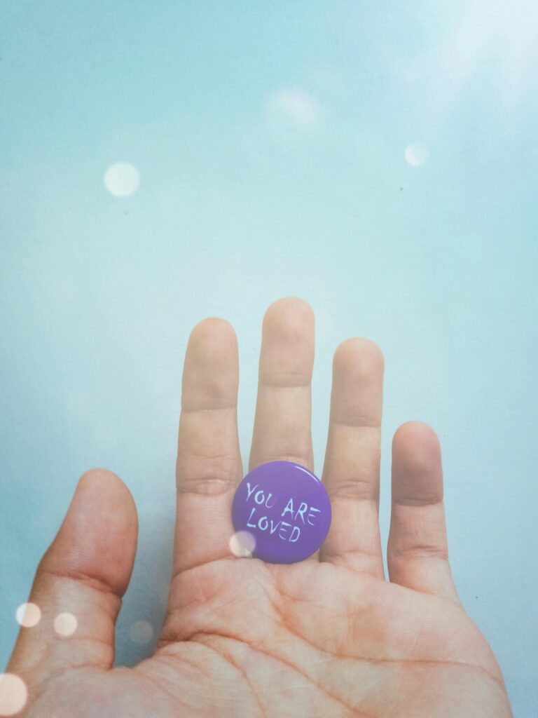 Close-up of a hand holding a purple pin with 'You Are Loved' message on a bright background.