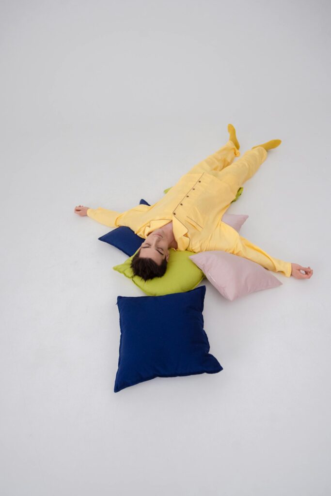 A man in yellow pajamas sleeps on colorful pillows, set against a white background.