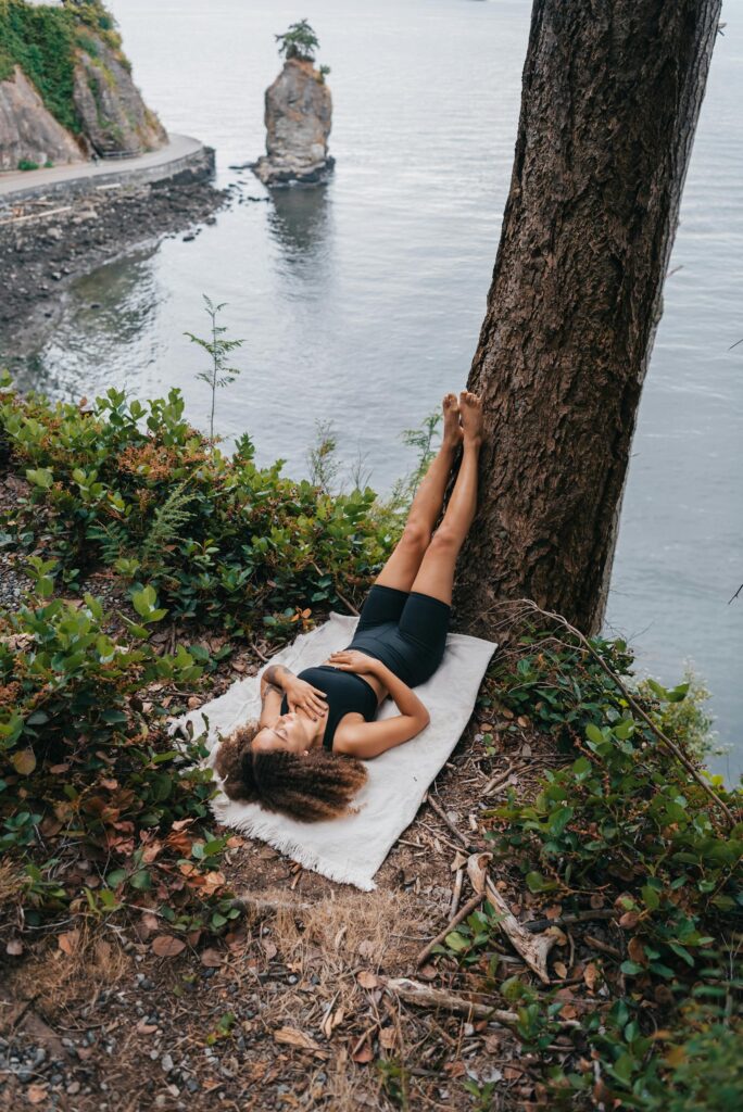 A woman in activewear relaxes by the water's edge with her legs on a tree, embracing tranquility.