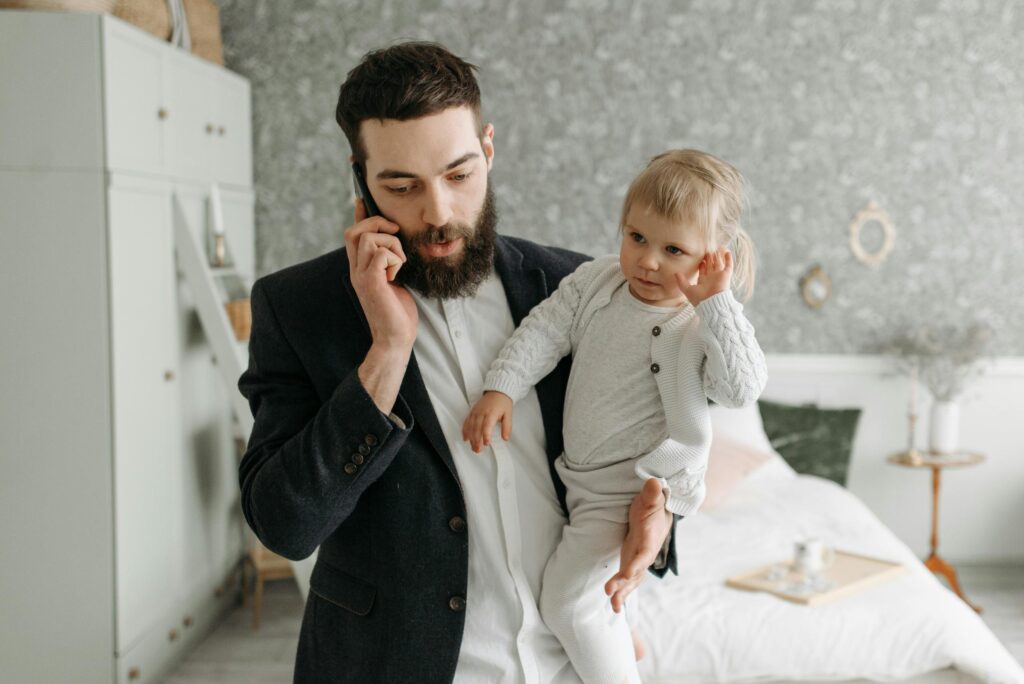 A bearded father talks on the phone while holding his baby in a cozy bedroom setting.